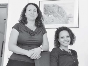 Dr. Jessica Vance Roitman (left) and PhD student Stacey Mac Donald of the Royal Netherlands Institute of Southeast Asian and Caribbean Studies KITLV in Leiden. In the background a historic photo of Saba’s Fort Landing before the harbour was built. (Suzanne Koelega photo)